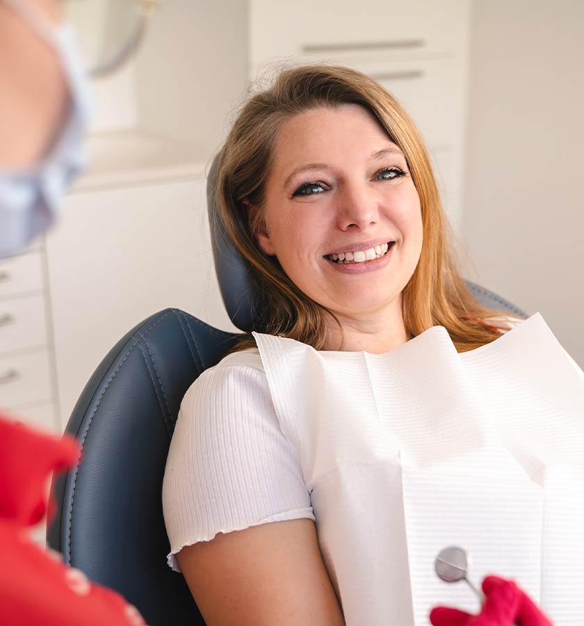 Lächelnde Patientin auf Behandlungsstuhl mit umgehängter weißer Dentalserviette im Gespräch mit Behandler bei MED:SMILE.