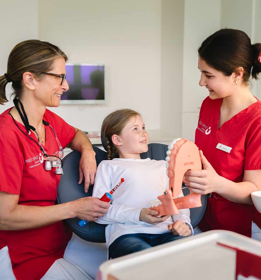 Lächelndes Mädchen auf Behandlungsstuhl bei der Zahnputzschulung zwischen ZÄ Wussidlo mit elektrischer Zahnbürste in der rechten Hand und MFA mit großem Modell eines Gebisses in der linken Hand. 