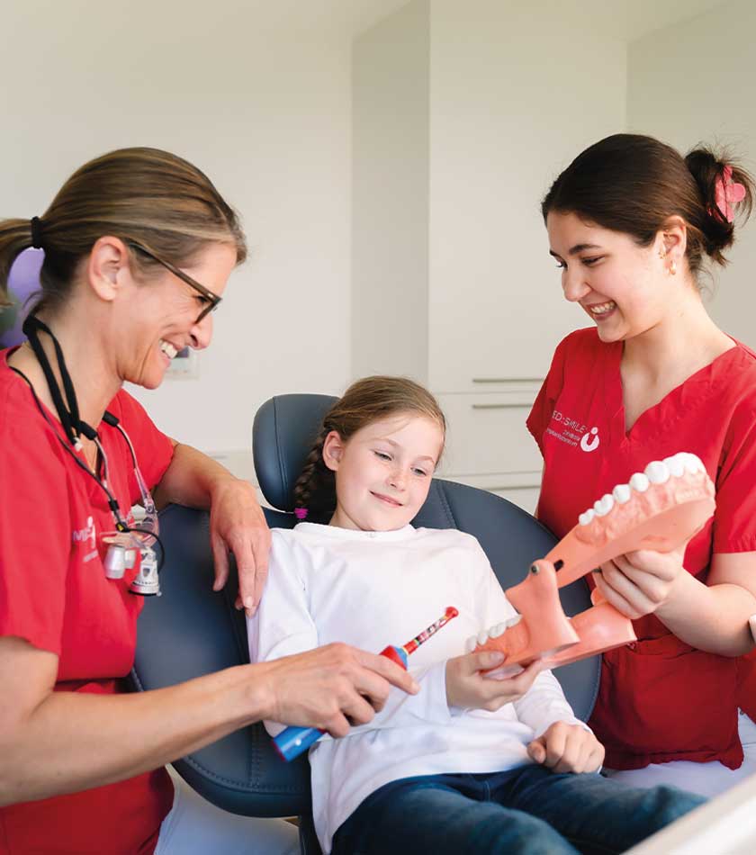 Kinderzahnärztin Julia Wossidlo und Mitarbeiterin erläutern junger Patientin Zahnputztechnik mit Zahnbürste und großem Gebissmodell