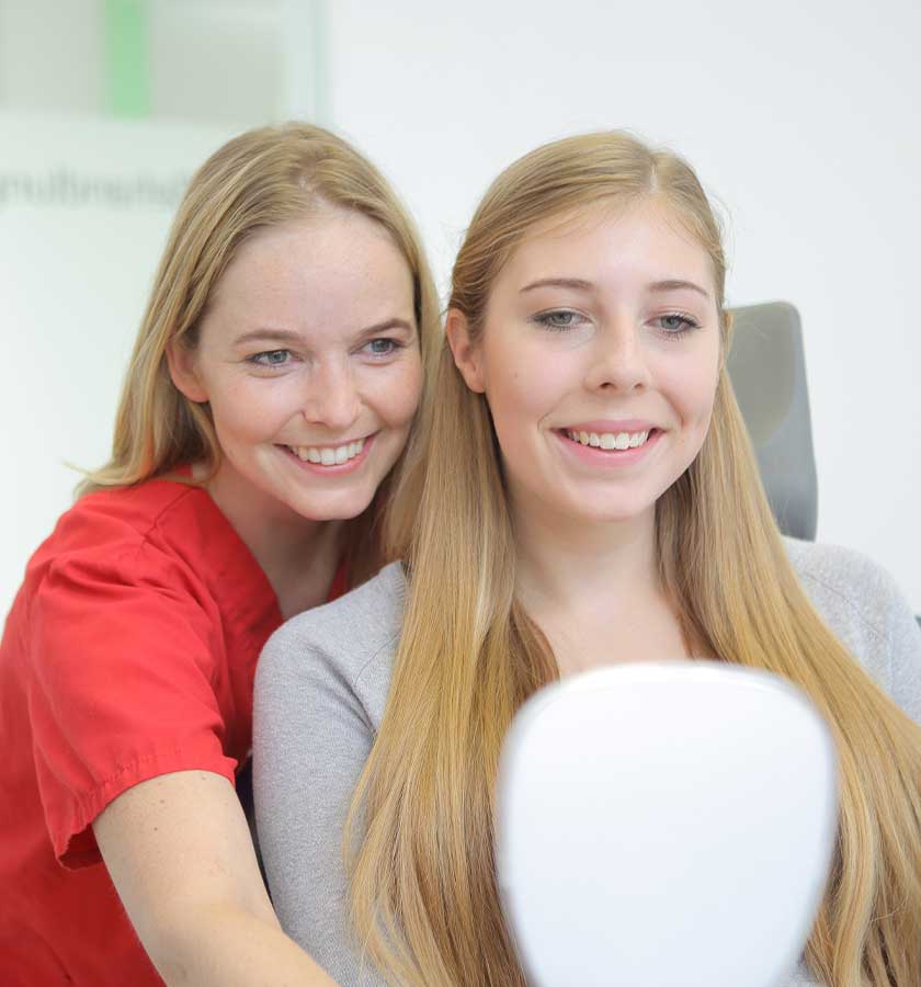 Lächelnde Patientin nach Bleaching beim Blick in den Spiegel, den MFA neben ihr für Sie hält.