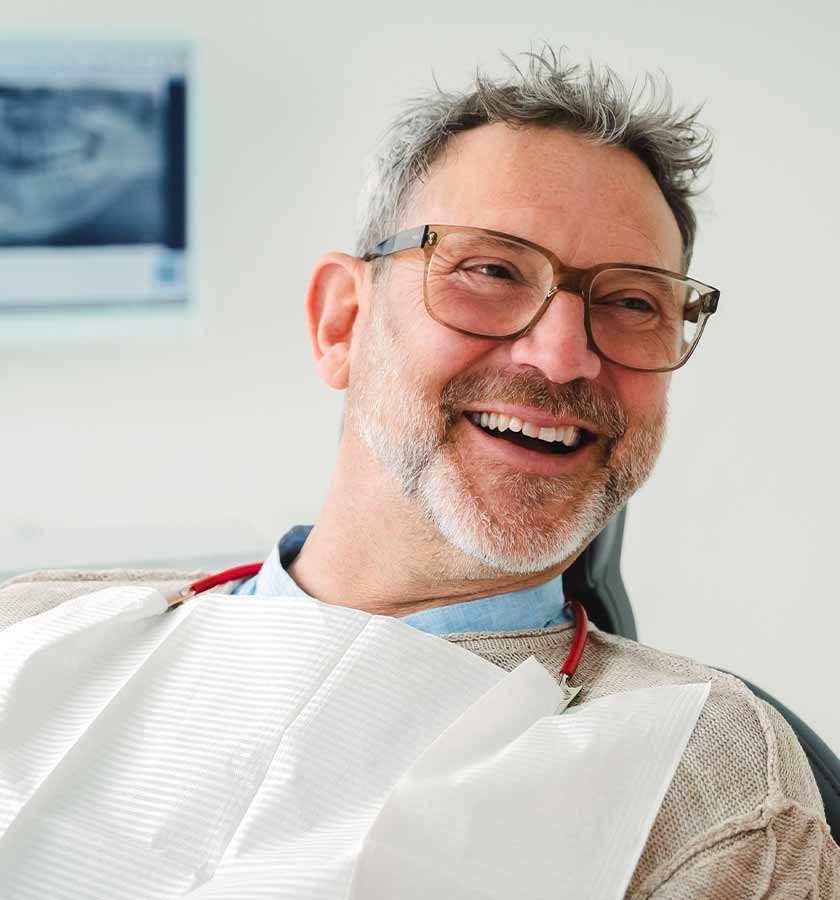 Lachender Patient mit Brille und Bart auf Behandlungsstuhl mit umgehängter weißer Dentalserviette.
