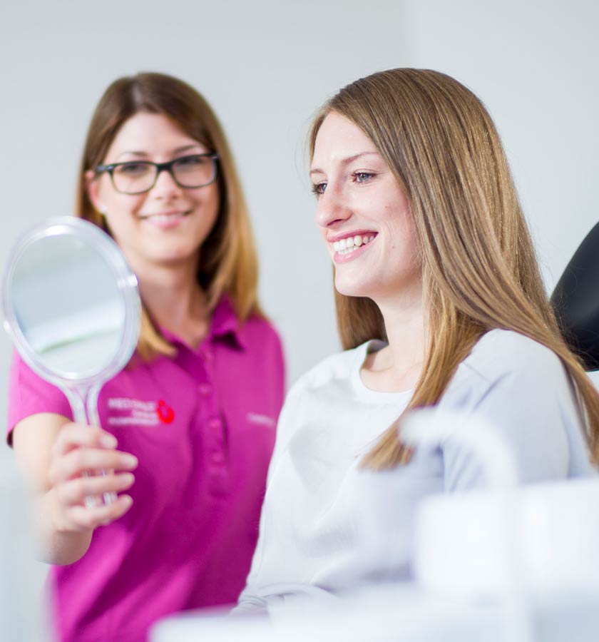 Lächelnde Patientin auf Behandlungsstuhl beim Blick in einen Handspiegel, den ihr eine lächelnde MFA hält.