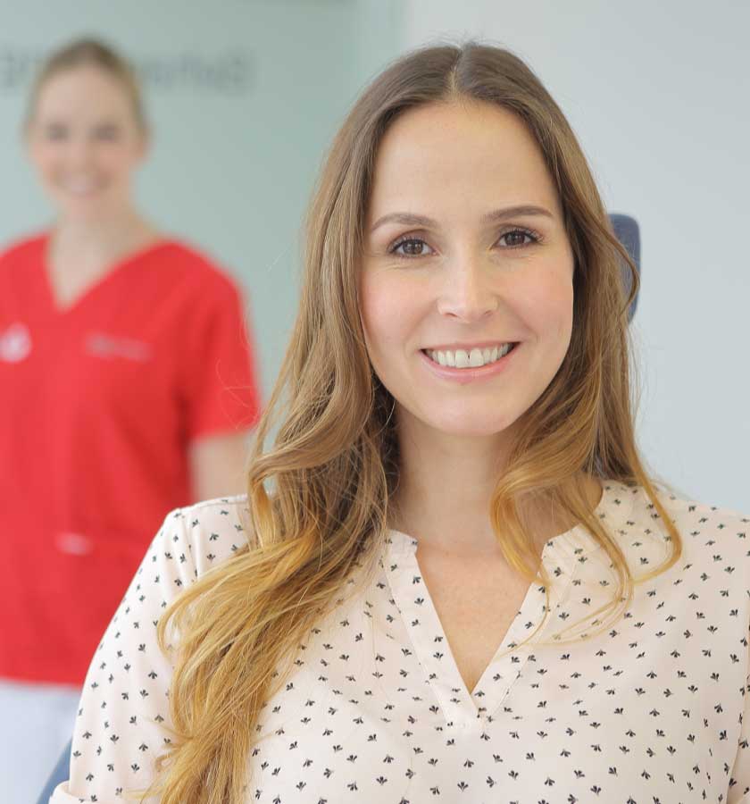 Blonde lächelnde Patientin auf Behandlungsstuhl mit MFA von MED:SMILE im Hintergrund.