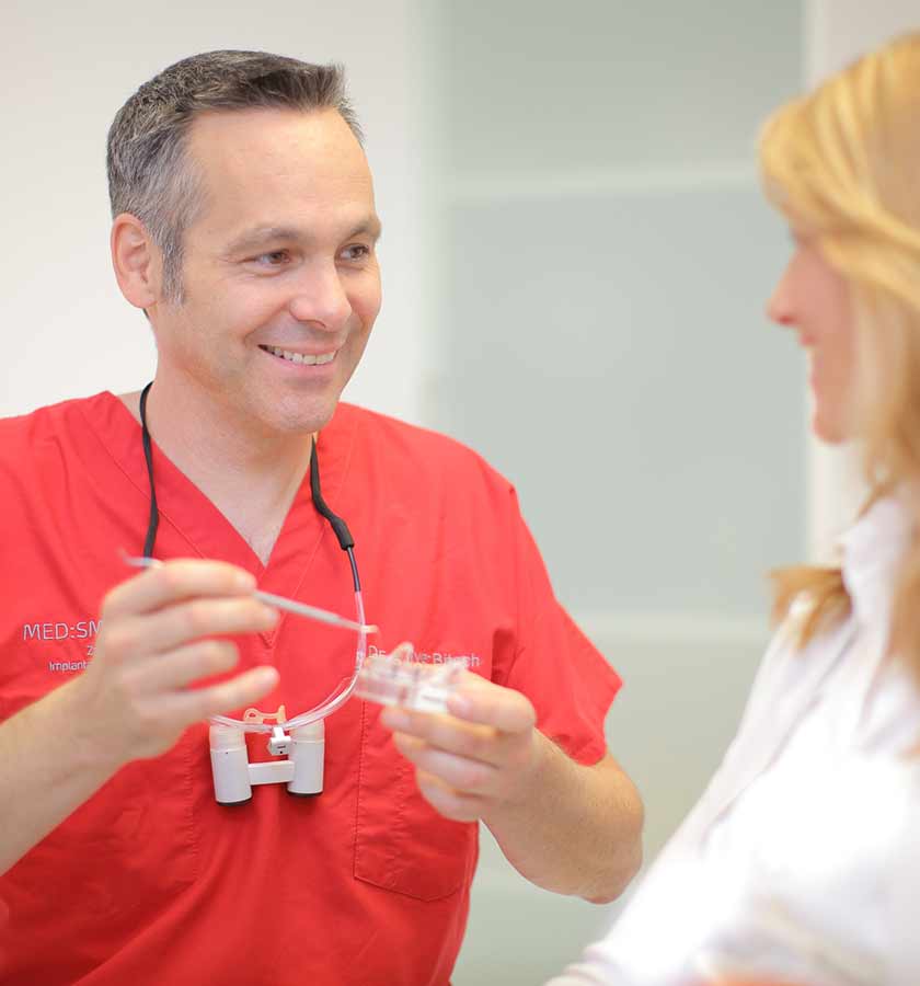 Dr. Bitsch Lupenbrille um den Hals und Dentalinstrument und transparentem Gebissmodell in den Händen im Gespräch mit Patientin.