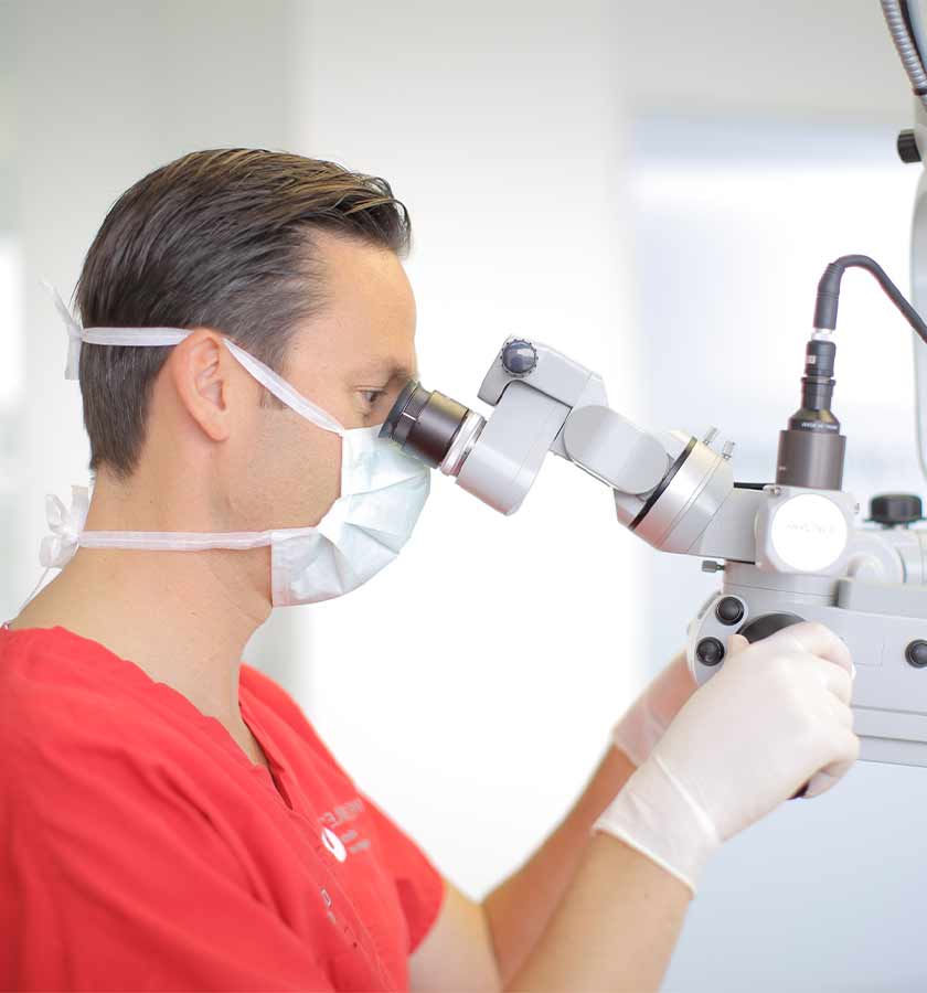 Seitenansicht von Dr. Bitsch mit Mundschutz beim Blick in ein OP-Mikroskop. 