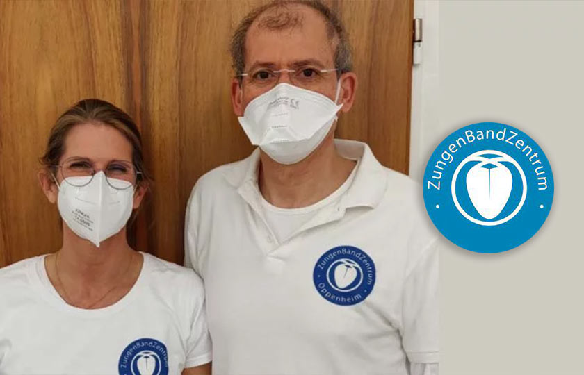 Zahnärztin Julia Wossidlo mit Mundschutz neben Kollegen mit Mundschutz neben weiß-blauem Logo des Zungenbandzentrums Oppenheim