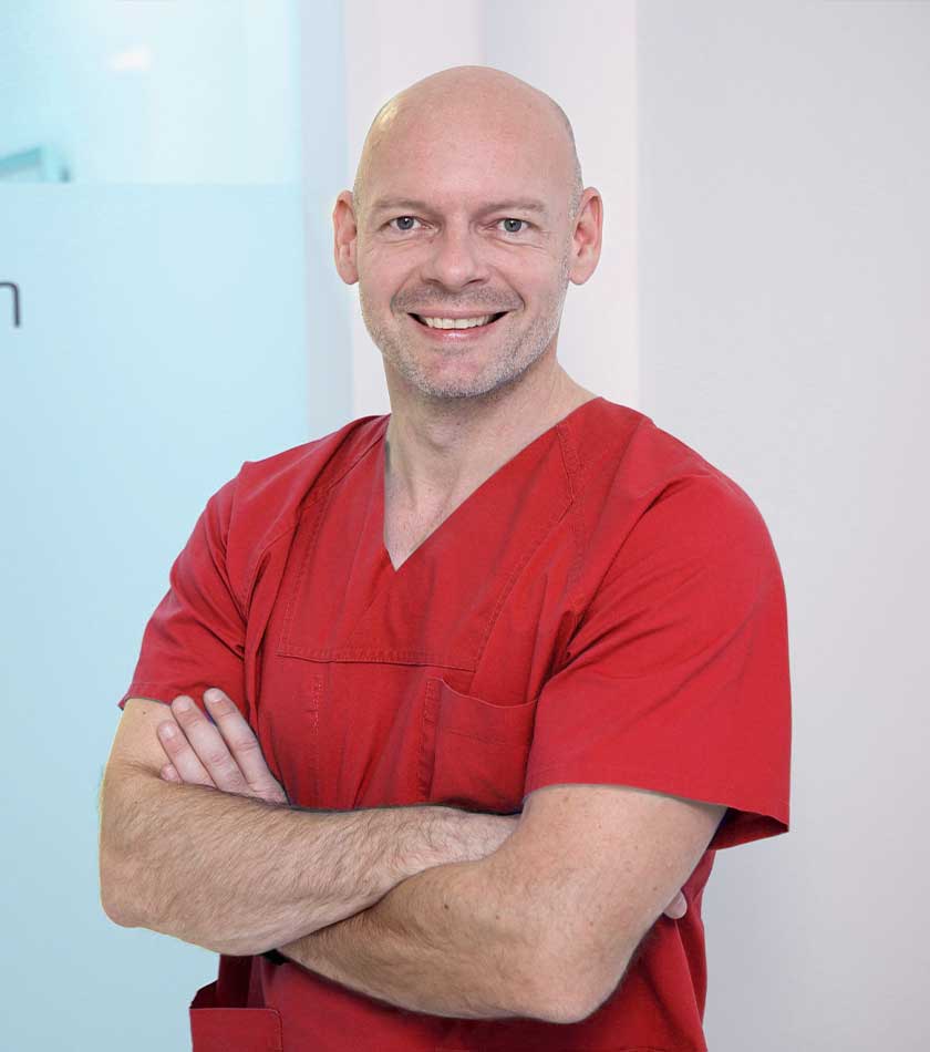Anästhesist Dr. Georg Grimm mit rotem Schlupfkasack vor Glastür