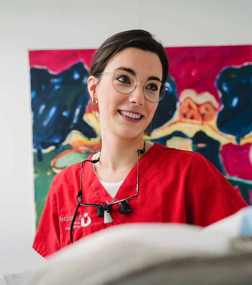 Zahnärztin Marie-Claire Schäfers mit Lupenbrille um den Hals vor buntem Gemälde im Gespräch