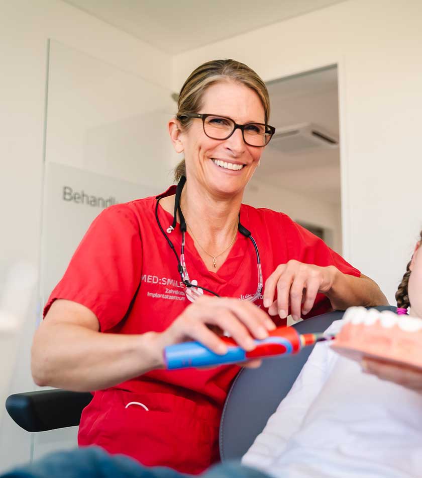 Sitzende Zahnärztin Julia Wossidlo mit elektrischer Zahnbürste in der Hand im Gespräch mit einer jungen Patientin, die auf dem Behandlungsstuhl sitzt